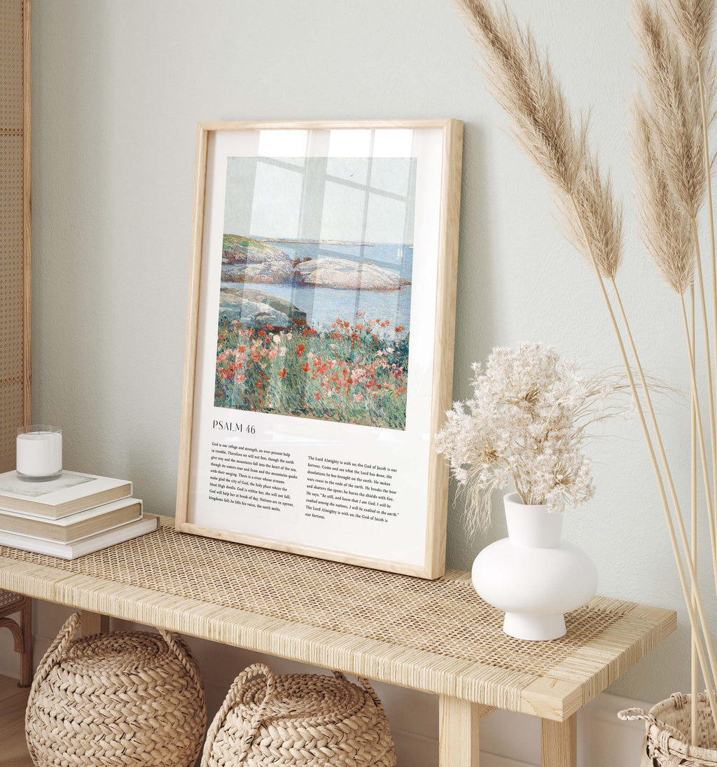 A framed art print featuring Psalm 24 and a coastal landscape with flowers and water, styled on a woven console table with books, a candle, a white vase of dried flowers, and pampas grass against a pale green wall.