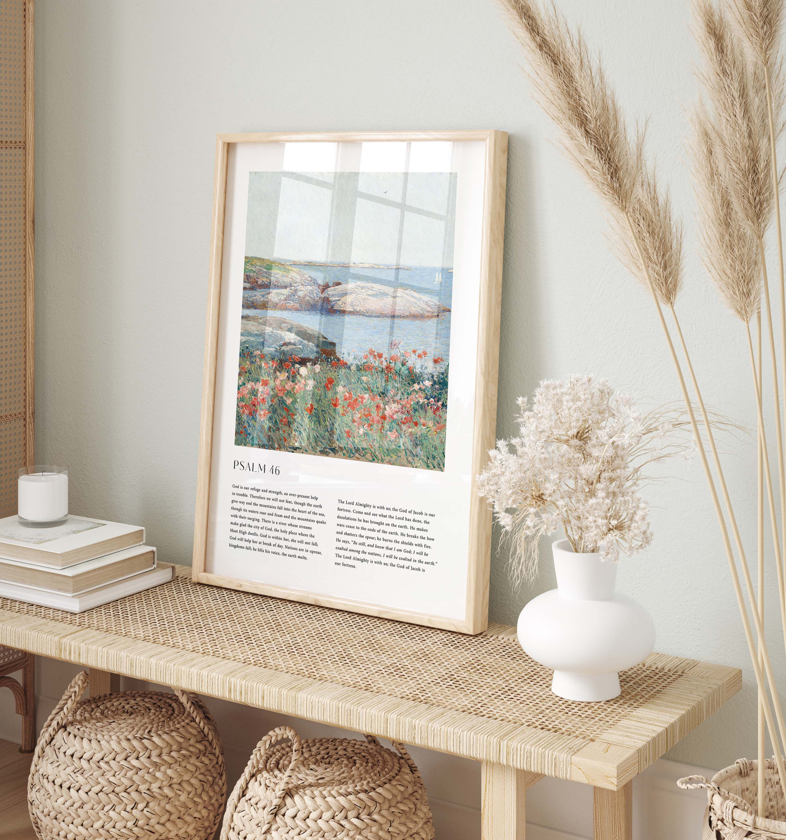 A framed art print featuring Psalm 24 and a coastal landscape with flowers and water, styled on a woven console table with books, a candle, a white vase of dried flowers, and pampas grass against a pale green wall.